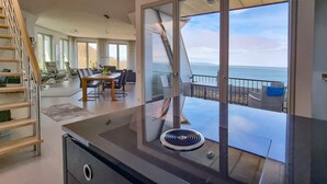 Une cuisine moderne dotée d’un grand îlot central, d’un coin repas avec une table et des chaises, et offrant une vue sur l’océan grâce à de grandes fenêtres.