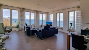 Une pièce spacieuse dotée de grandes fenêtres offrant une vue sur la mer, d’un coin salon confortable, d’un bureau et d’une télévision.