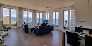 Quarto espaçoso com grandes janelas com vista para o mar, uma área de estar confortável, uma escrivaninha e uma televisão.
