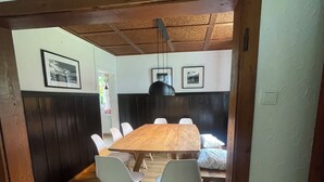 A dining area with a wooden table and white chairs, a black pendant light, and framed pictures on the walls.