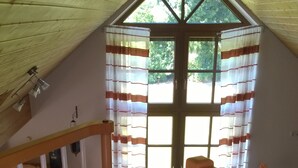 Ein Zimmer mit schrägen Wänden, einer Holztreppe, einem Essbereich mit Tisch und Stühlen sowie einem Fenster mit gestreifter Gardine.