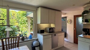 Una cocina compacta con armarios blancos, un microondas y un refrigerador. Hay una mesa de comedor con sillas y una ventana con vistas a zonas verdes.