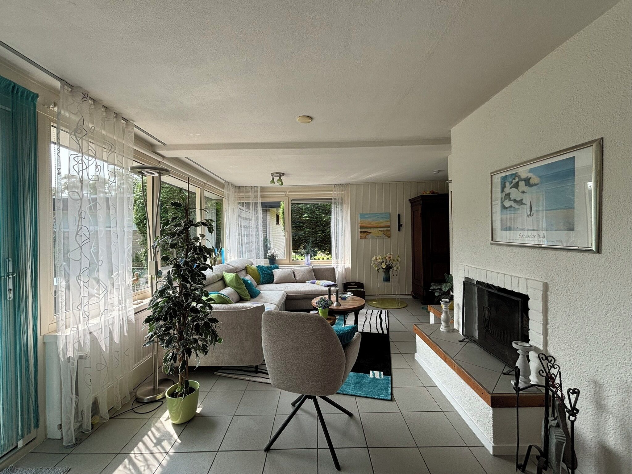 A spacious living room with a large sofa, a coffee table, and a fireplace. There are several potted plants and a painting on the wall.