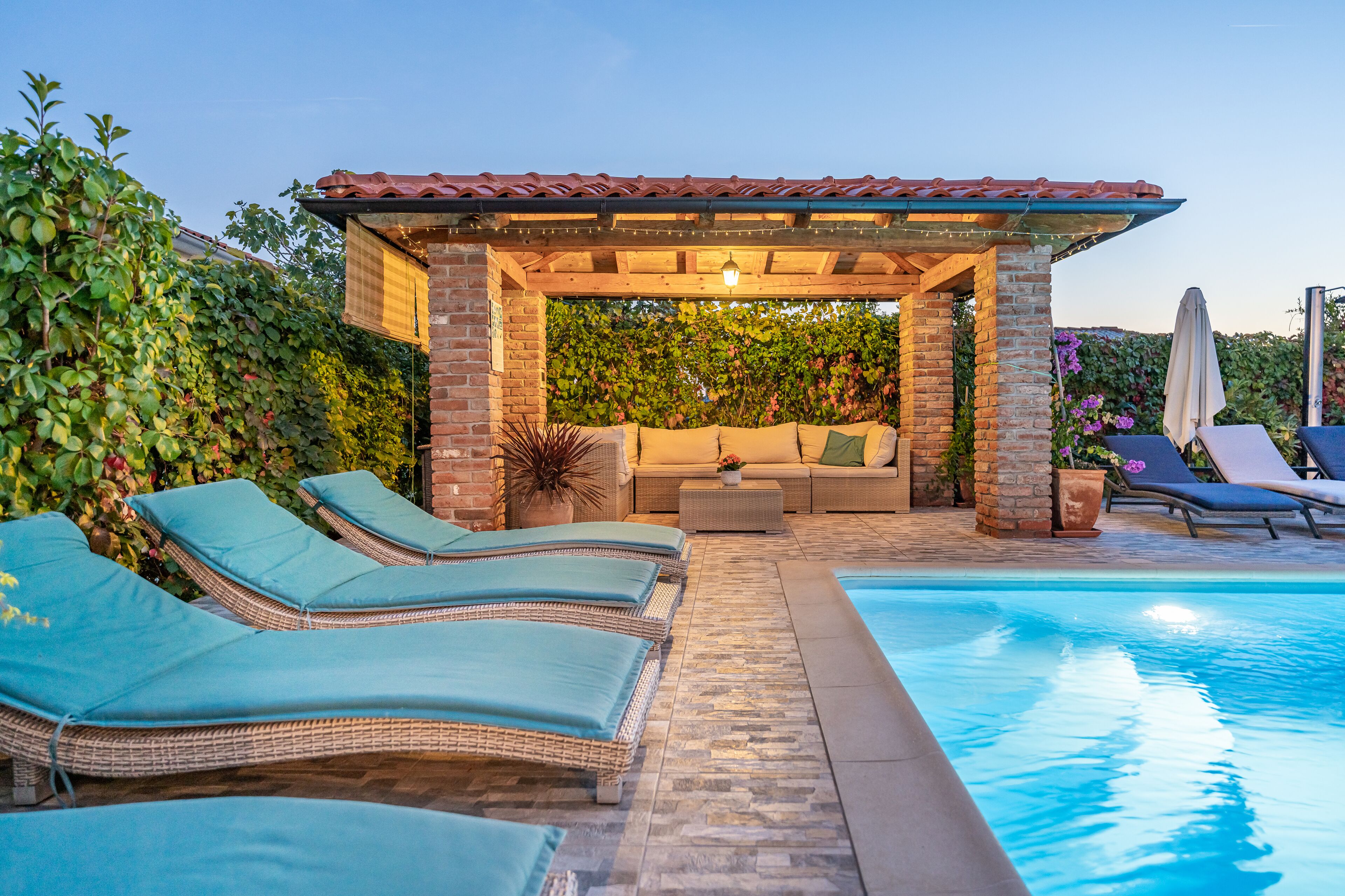 A pool area with lounge chairs, a covered seating area, and a brick wall.