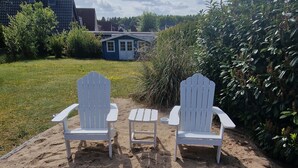 Two white Adirondack chairs with a small table in the sand, surrounded by greenery and a house in the background. - Haus Lena - Holiday near the Baltic Sea including free Wi-Fi (V-DSL) (Pansdorf)