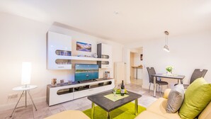 A modern living room with a flat-screen TV, a dining area with a table and chairs, and a sofa with cushions.