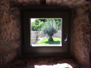 A room with a stone wall and a window overlooking a garden with a tree and a stone wall.