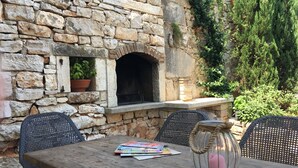 Un muro de piedra con chimenea integrada, mobiliario exterior con sillas de mimbre y una mesa de madera con revistas y un frasco decorativo.