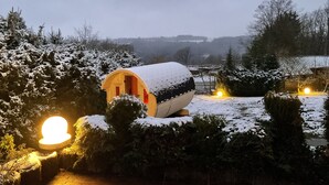 Ein verschneiter Holzbau mit Tür und Fenstern, umgeben von schneebedecktem Gebüsch und Bäumen, unter einem bewölkten Himmel.