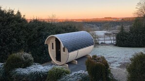 Eine zylindrische Holzkonstruktion mit sechseckigem Dach, umgeben von frostbedeckten Büschen und Bäumen, vor einer Kulisse eines Sonnenuntergangs über einer bewaldeten Landschaft.