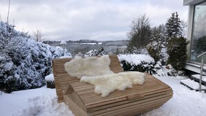 Eine hölzerne Bank mit weißem Fellüberwurf, umgeben von schneebedeckten Bäumen und Blick auf eine ferne Landschaft.