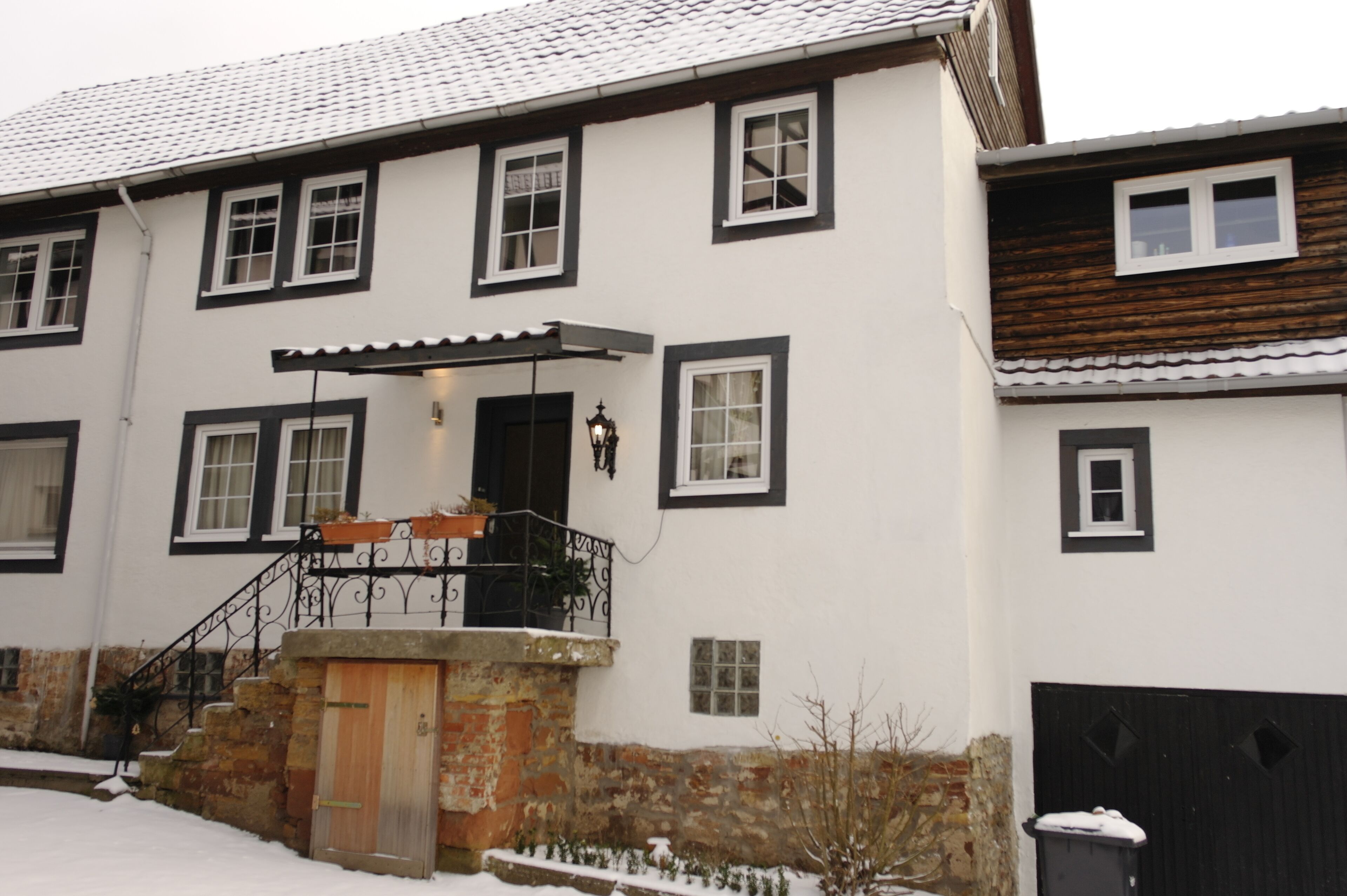 A two-story building with a stone foundation, a wooden door, and a black metal staircase leading to a porch with a roof.