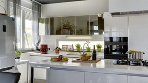 Une cuisine moderne avec des meubles blancs, un îlot central, un four encastré et une grande fenêtre avec des rideaux.