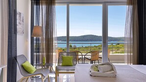 Une chambre d’hôtel avec un lit, une chaise dotée d’un coussin vert, une petite table avec une lampe, et une vue sur la mer et les collines.