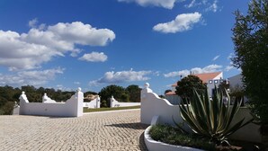 Une allée dallée, bordée de murs blancs, mène à un bâtiment dont le toit est recouvert de tuiles rouges, par un chemin sinueux.