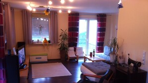 Una sala de estar acogedora con un sofá, un sillón y una mesita. Hay un televisor, una ventana con cortinas y una planta.