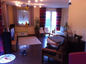 Uma sala de estar aconchegante com sofá, poltrona e uma mesinha. Há uma televisão, uma janela com cortinas e uma planta.