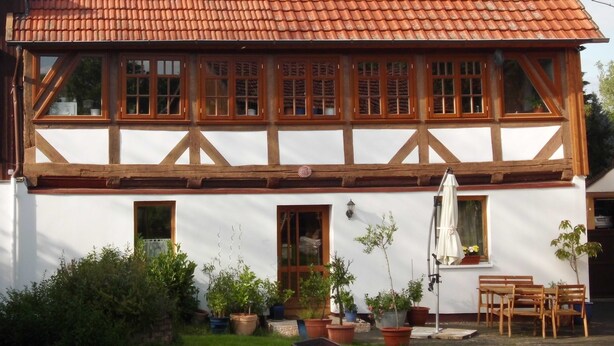 Ein traditionelles Fachwerkhäuschen mit Ziegeldach, zahlreichen Fenstern und einem kleinen Garten mit Blumentöpfen und Gartenmöbeln.
