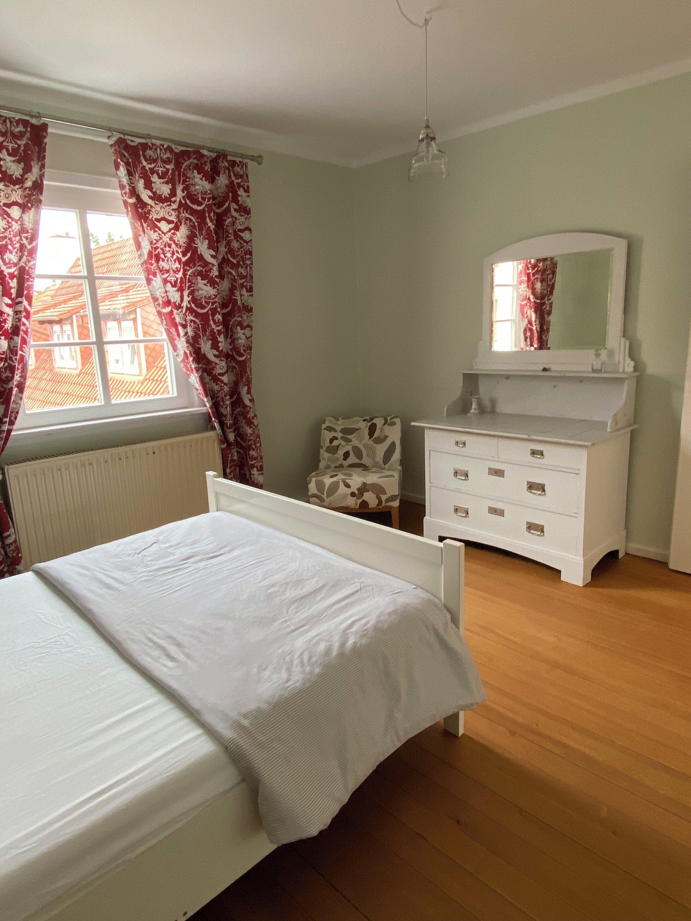 Une chambre à coucher comprenant un lit, une commode, une chaise, une fenêtre avec des rideaux et un luminaire suspendu.