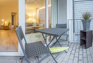 Varanda com duas cadeiras dobráveis e uma pequena mesa, um vaso de planta e vista para um espaço de estar interno.