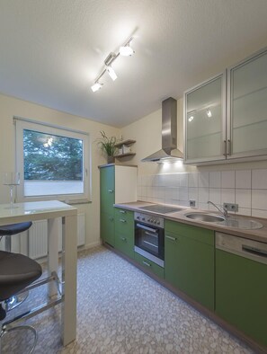 Uma cozinha moderna com armários verdes, coifa de aço inoxidável, janela com vista para árvores e um banquinho de bar.