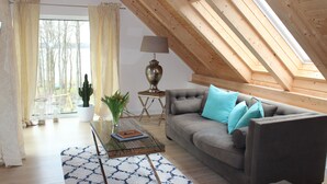 Uma sala de estar aconchegante no estilo loft, com teto inclinado, sofá cinza com almofadas azuis, uma mesa de centro de vidro e um abajur.