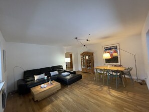 Ein Wohnzimmer mit einer schwarzen Ledersofa, einem Holztisch und einem Esstisch mit Stühlen. Außerdem ist ein Holzschrank und eine Lampe im Raum.