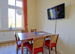 Ein Essbereich mit einem Holztisch und vier Stühlen, einem an der Wand montierten Fernseher und einem Fenster mit Vorhängen.