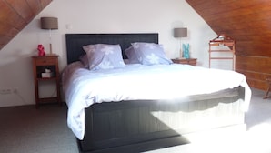Ein Schlafzimmer mit geneigter Decke, einem Bett mit weißer Bettwäsche, zwei Nachttischlampen, einem Nachttisch und einem kleinen Regal mit einer Lampe und einer Figur.