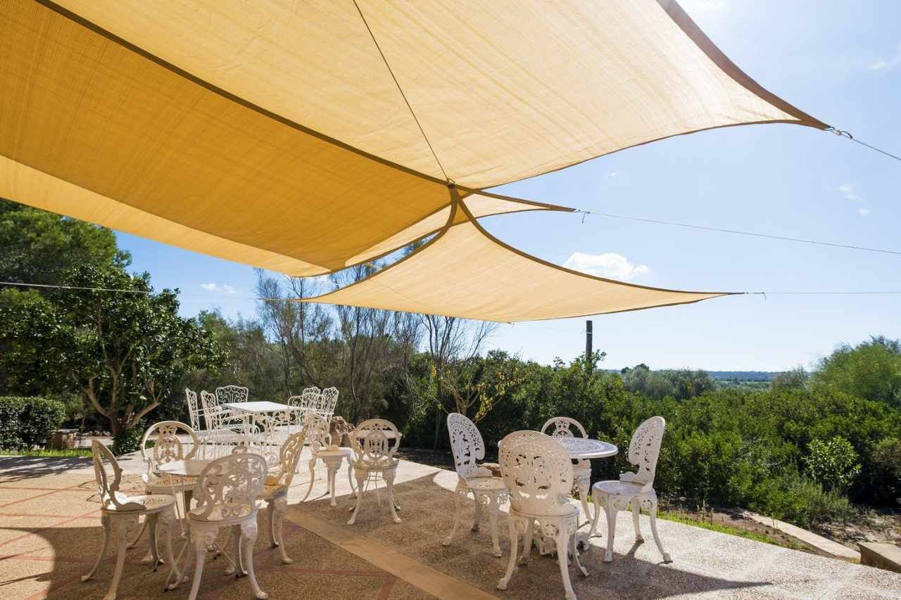 Un espace extérieur aménagé avec des chaises et des tables en métal blanc, placé sous une grande tonnelle beige, et entouré de verdure et d’arbres.
