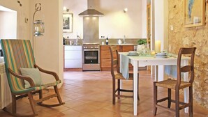 A dining area with a table set for two, wooden chairs, a rocking chair, a kitchen in the background, and a stone wall.