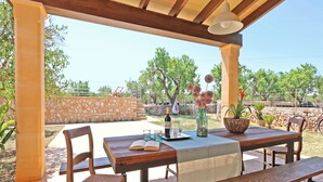 An outdoor patio with a wooden table and chairs, a bottle of wine, a glass pitcher, and a vase with flowers.