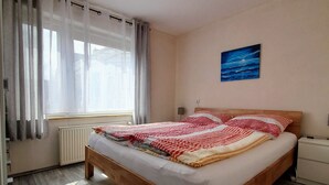 Makuuhuoneessa on puinen sänky, patteri, ikkuna, jossa on verhot, ja seinällä on taulu.