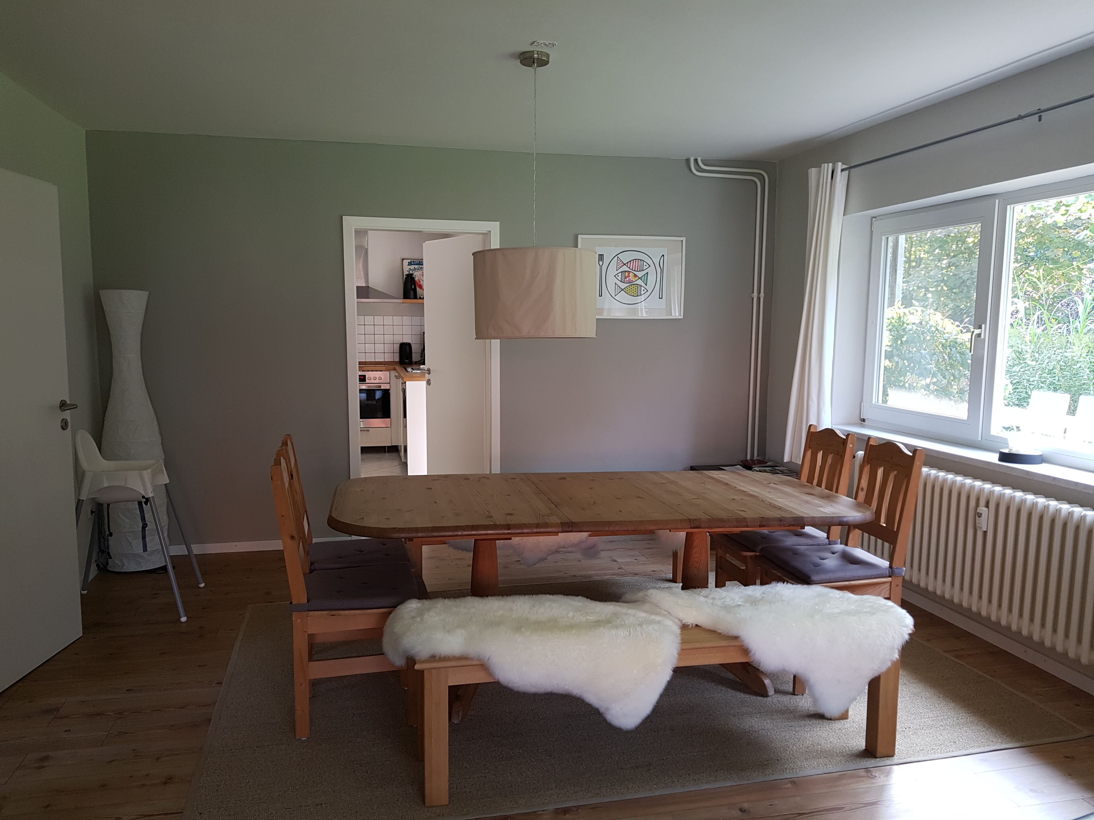 Un comedor con una mesa y sillas de madera, un cubrezonas de piel en una silla, una cocina al fondo y una ventana con vista a follaje.
