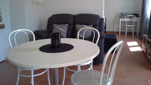 Ein Wohnbereich mit einem runden Tisch, weißen Stühlen, einem schwarzen Sofa mit gemusterten Kissen und einem kleinen weißen Beistelltisch.