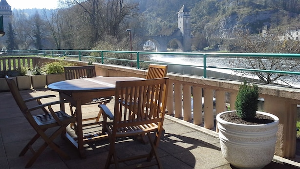 Eine Terrasse mit Tisch und Stühlen, mit Blick auf einen Fluss, eine Brücke und Bäume im Hintergrund.