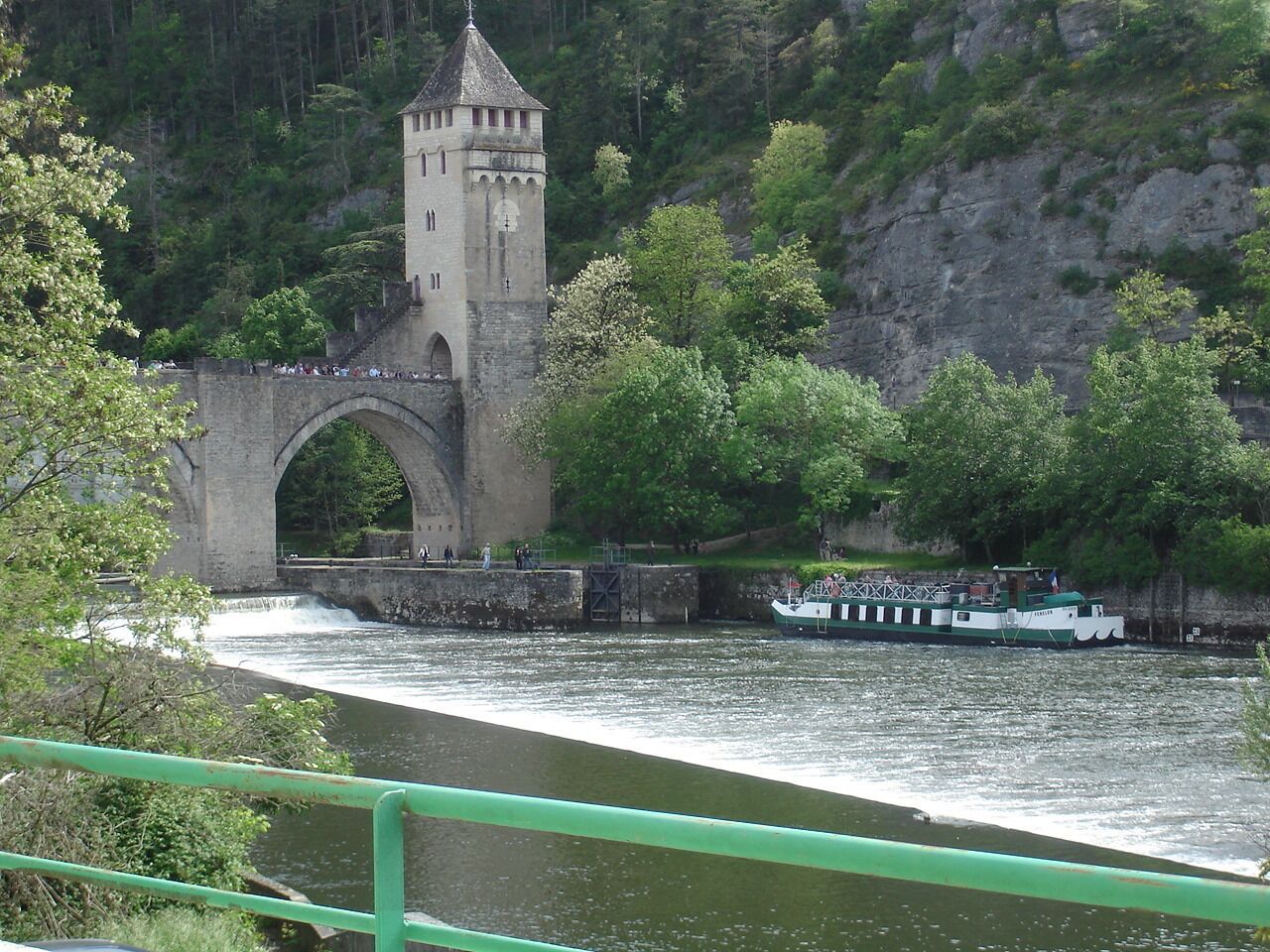A stone bridge with a tower, a boat on the river, and greenery around.