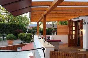 A covered outdoor area with a wooden pergola, seating, and a view of the surrounding garden and buildings.