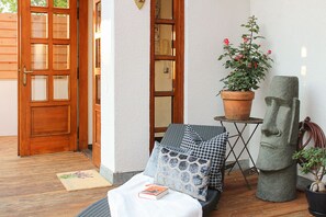 A wooden door, a potted plant with pink flowers, a stone sculpture, and a small table with a patterned cushion and a book.