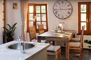 Een eetruimte met een houten tafel, stoelen, een grote wandklok en een raam met gordijnen.