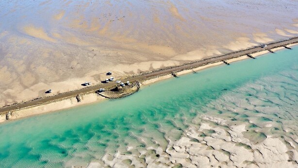An aerial view of a beach with a pier extending into turquoise waters, a sandy shore, and a few parked vehicles.