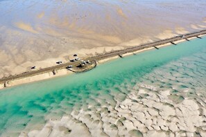 An aerial view of a beach with a pier extending into turquoise waters, a sandy shore, and a few parked vehicles.