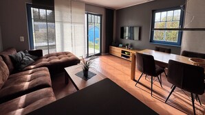 A modern living room with a sofa, a coffee table, a dining table with chairs, a TV mounted on the wall, and large windows offering a view of the outdoors.