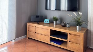 A modern living room with a flat-screen TV mounted on the wall, a wooden entertainment unit with a speaker, a small plant, and a calendar.