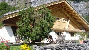 Eine Holzhütte mit geneigtem Dach, einer Veranda mit Holzstühlen und im Hintergrund ein Blick auf eine Felswand und einen Wasserfall.