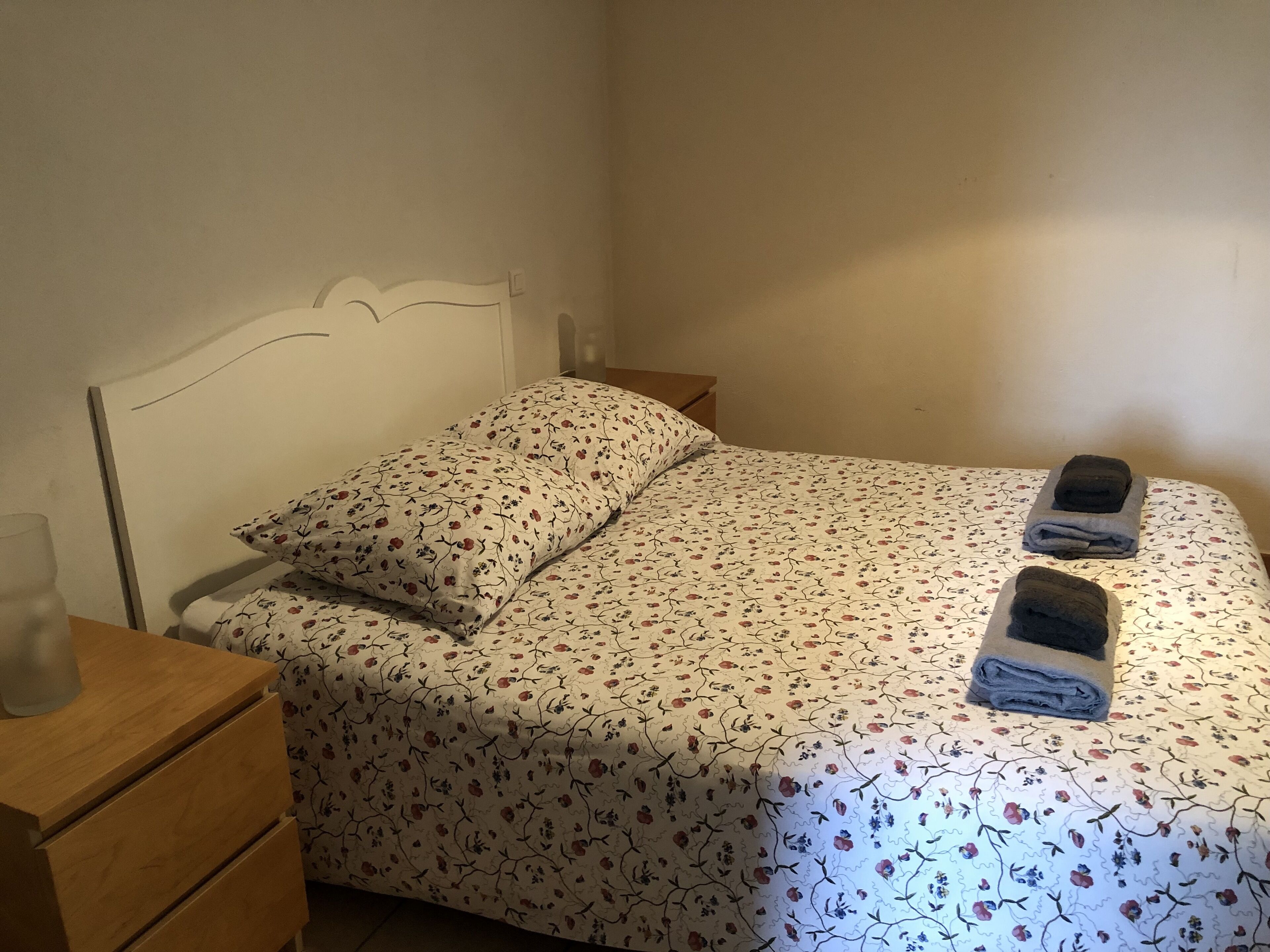 Een net opgemaakt bed met bloemenbeddels, een houten nachtkastje met een glas en twee handdoeken op het bed.