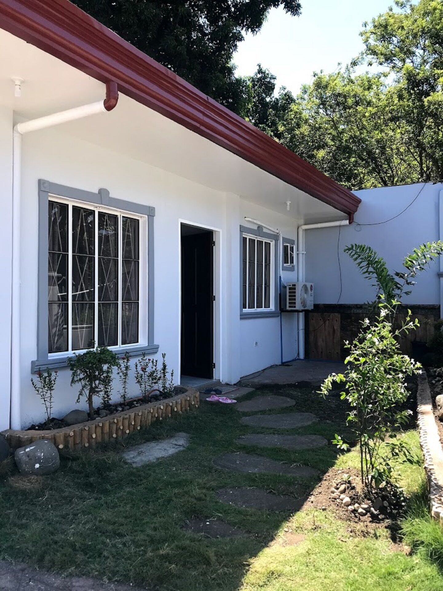 A single-story house with a red roof, white exterior walls, and a small front yard with a garden bed and a few plants.