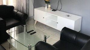 Una sala de estar moderna con una mesa de centro de vidrio, sofás de cuero negro, un mueble de televisión blanco con una televisión de pantalla plana y un florero con flores.
