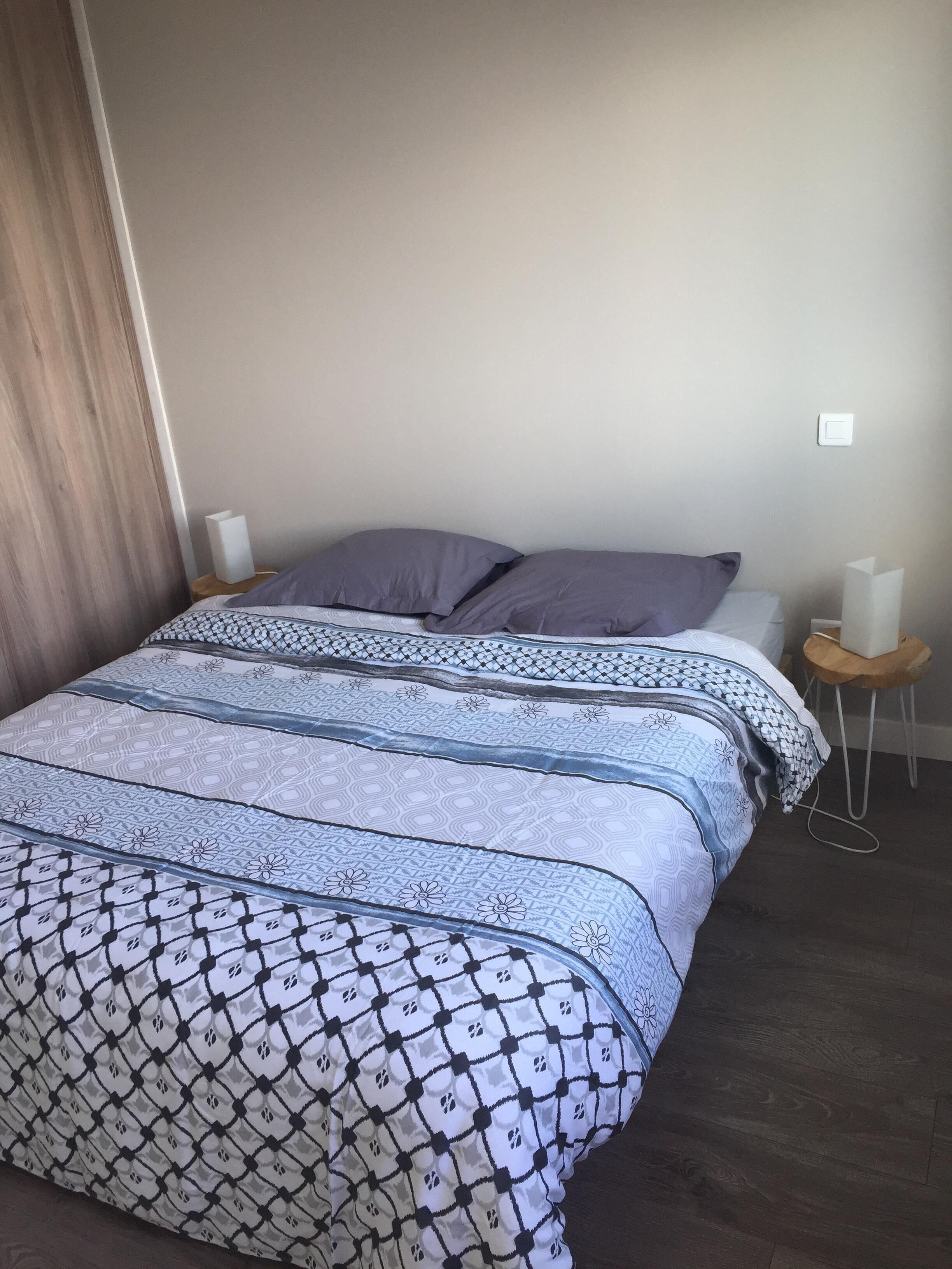 Una cama bien tendida con una colcha estampada y almohadas, dos mesitas de noche con lámparas, y suelo de madera.
