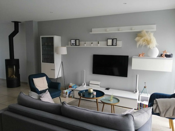 A modern living room with a grey sofa, a blue armchair, a black fireplace, a flat-screen TV, and a white shelf with decorative items.
